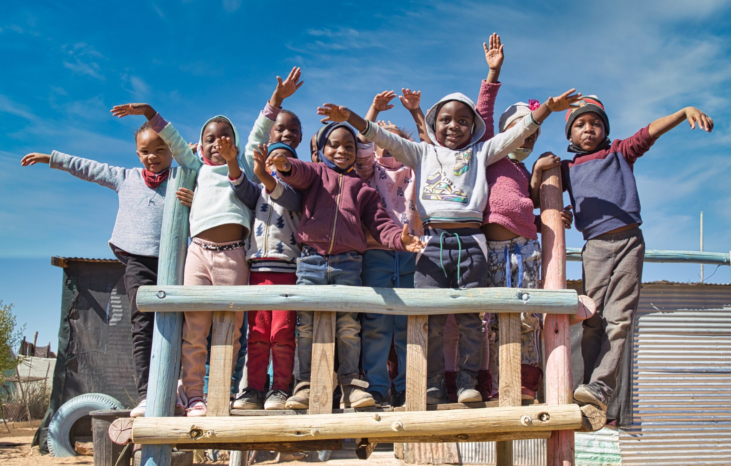 A group of young children waving happily while standing on a wooden platform.